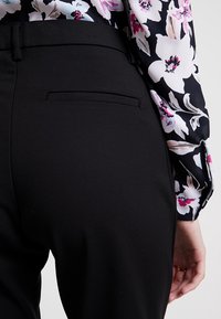 Person klädd i svarta skräddarsydda byxor med bakficka och en blommig långärmad blus i rosa, vitt och svart.