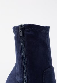 Acercamiento a una bota de ante azul marino de tobillo con cremallera lateral visible y costura detallada en la parte superior.