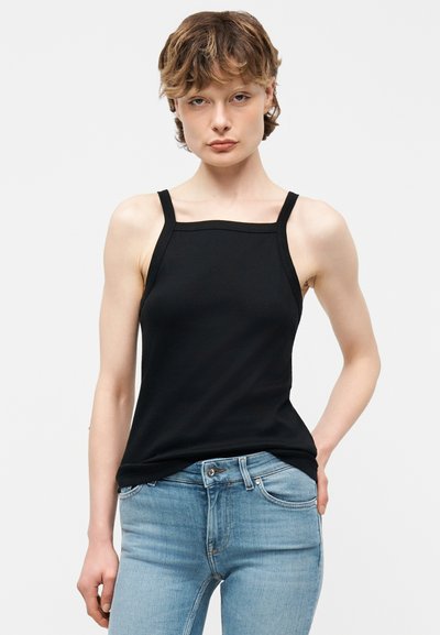 Νεαρό άτομο με κοντά σγουρά μαλλιά, ντυμένο με μαύρο αμάνικο τοπ και ανοιχτό μπλε τζιν, στέκεται μπροστά σε απλό λευκό φόντο.