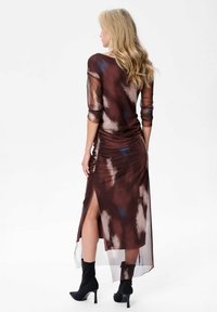 Robe transparente à motifs marron avec de longues manches et une fente sur le côté. Le tissu texturé présente des accents bleus et beiges. Associée à des bottines noires.