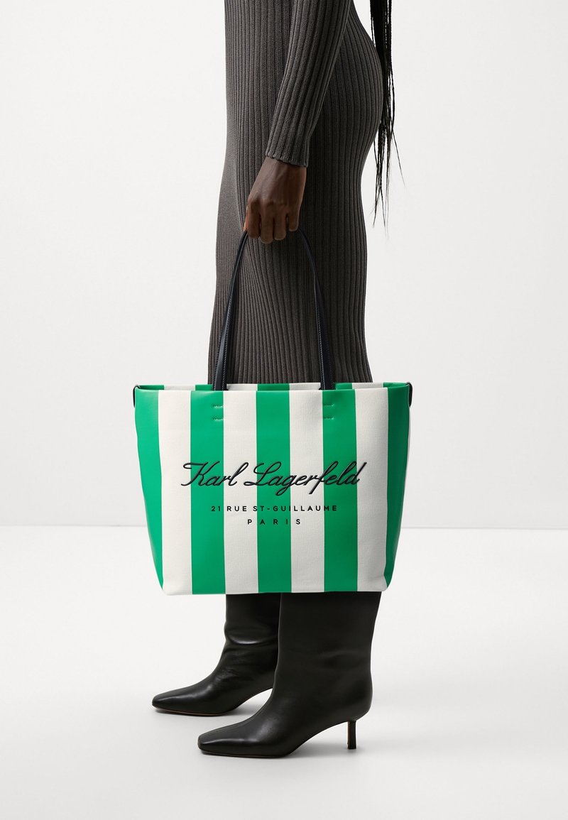 Femme portant une robe grise côtelée et des bottines noires à talons, tenant un sac fourre-tout rayé vert et blanc Karl Lagerfeld.