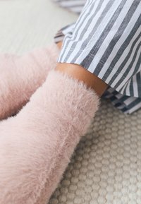 Zachte roze sokken bedekken de enkels, gecombineerd met gestreepte stof aan de zoom van de broek. Op de voorgrond ligt een gestructureerd vloerkleed in neutrale kleuren.