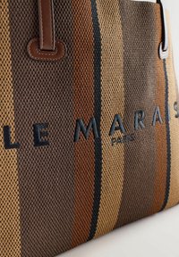 Tkaná taška na rameno s horizontálnymi pruhmi v odtieňoch hnedej, béžovej a čiernej. Dopĺňajú ju hnedé kožené rukoväte a vyšívaný text.