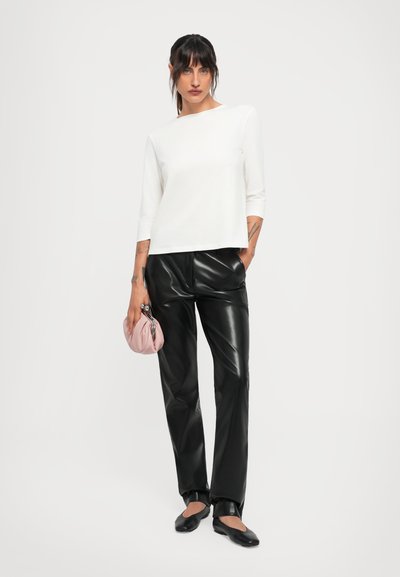Woman wearing a white long-sleeve top, black leather pants, black flat shoes, and holding a small pink clutch bag, standing against a white background.