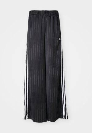 Pantalon large noir avec des rayures blanches verticales, taille élastique et trois rayures blanches sur le côté. Logo Adidas à l'avant.