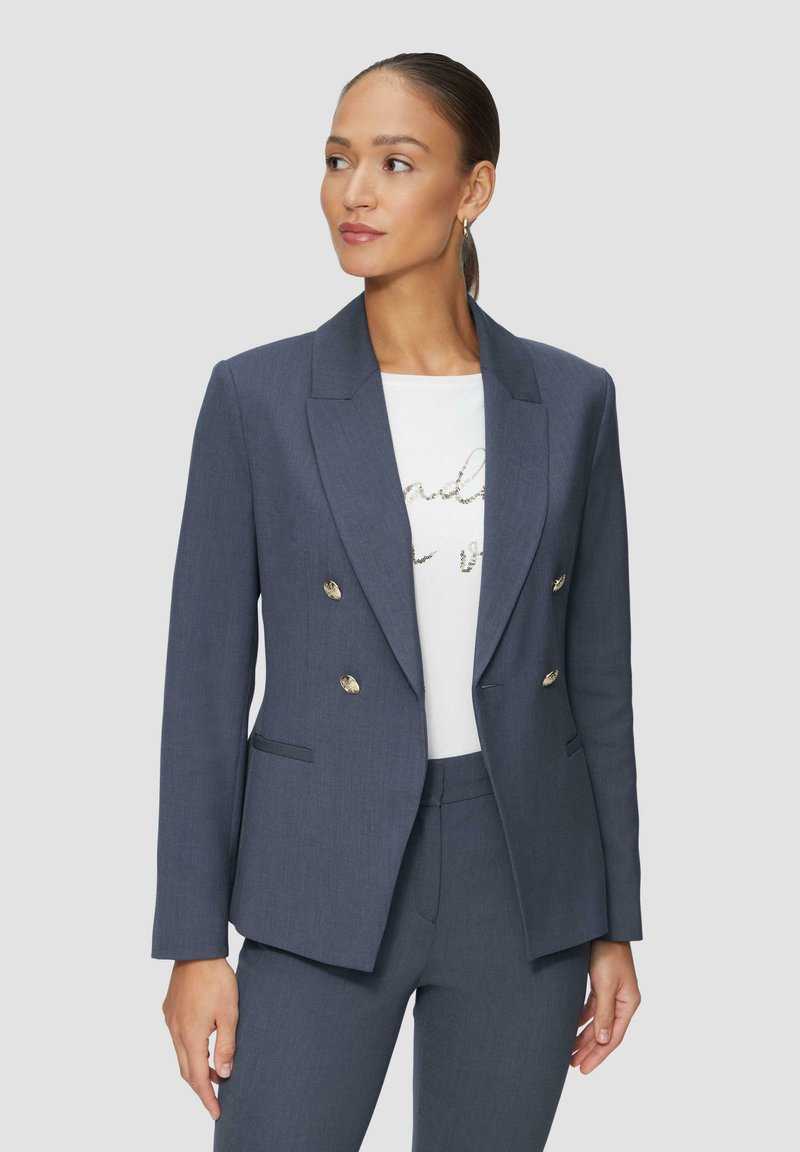 Blazer ajusté bleu foncé à double boutonnage avec des accents de boutons dorés, revers à cran et poches latérales, porté sur un tee-shirt graphique blanc.