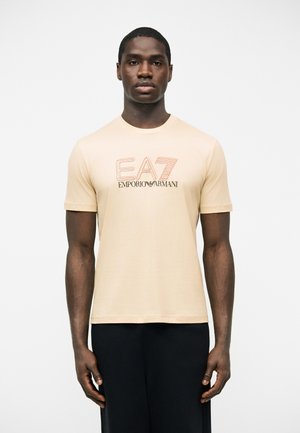 Homme portant un T-shirt beige EA7 Emporio Armani et un pantalon noir, debout devant un fond blanc uni, faisant face à l'avant.