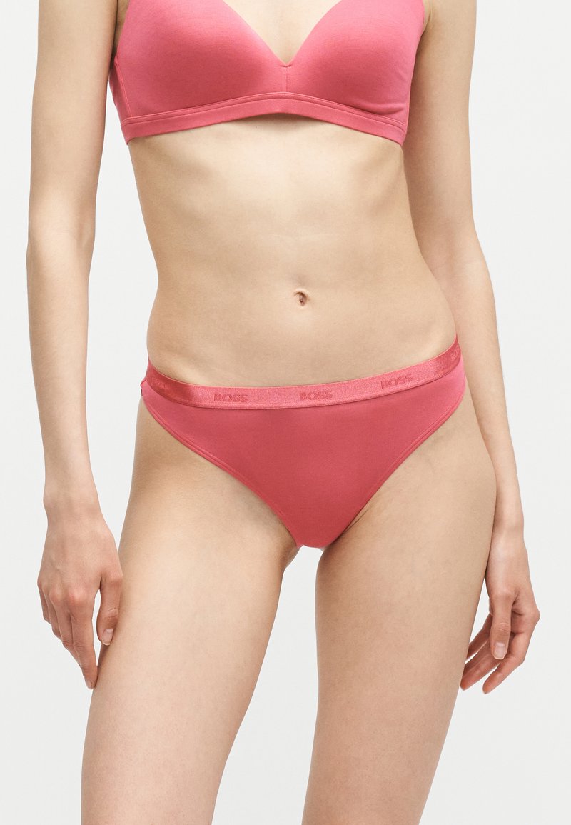Femme portant un soutien-gorge et une culotte bikini rose corail assortis avec le logo BOSS sur la ceinture, debout devant un fond clair uni.