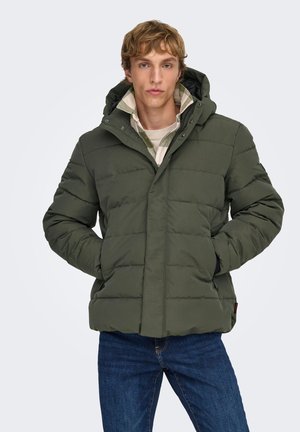 Jeune homme portant une veste matelassée à capuche verte par-dessus une chemise à carreaux claire et un t-shirt blanc, les mains dans les poches de la veste, debout devant un fond blanc.