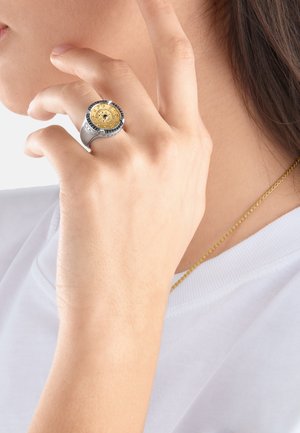 Main avec une bague à thème engrenage en or et argent touchant le visage, portant un haut blanc et un collier chaîne fine doré.