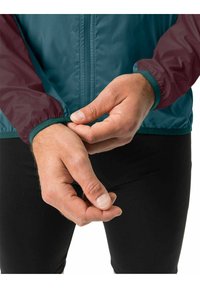 Giacca leggera bicolore con corpo teal e maniche bordeaux. Presenta polsini elasticizzati e chiusura con zip frontale. Progettata per la flessibilità.