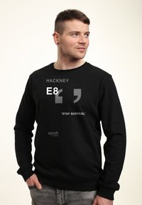 Felpa nera con collo tondo, caratterizzata da testo grigio: "HACKNEY E8" e "'STAY SURREAL'" sulla parte anteriore, realizzata in tessuto morbido.