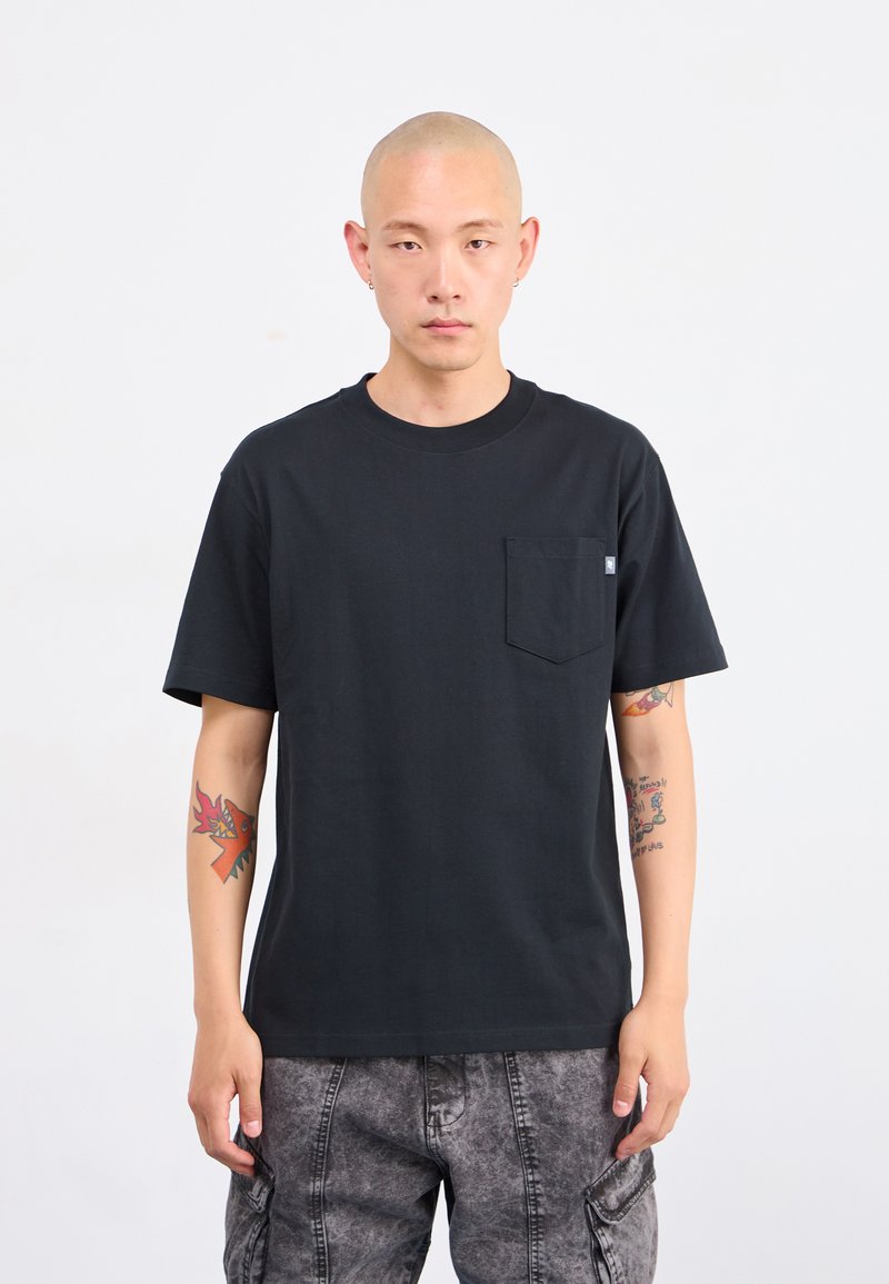 T-shirt en coton noir avec un col rond et des manches courtes, doté d'une poche sur la poitrine. Le tissu semble lisse avec une coupe décontractée.