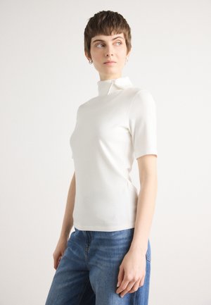 Witte top met korte mouwen, hoge hals en ritsdetail. Strak model met een gladde textuur, gecombineerd met een blauwe wijde jeans.