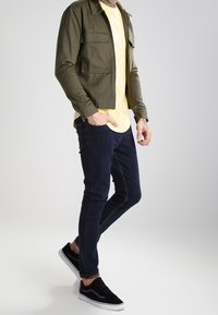 Hombre vestido con una chaqueta verde olivo, camiseta amarilla, jeans azul oscuro y zapatillas negras, de pie con una mano en el bolsillo, contra un fondo claro.