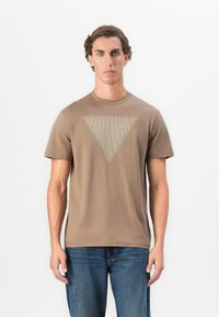 Joven con el cabello rizado y corto, vistiendo una camiseta marrón con un diseño de triángulo invertido a rayas y jeans azules, de pie frente a un fondo blanco.
