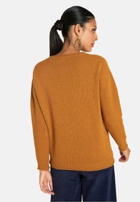 Knitpullover in verbranntem Orange mit Rundhalsausschnitt, weiten Ärmeln und geripptem Saum. Getragen mit dunkelblauen Cordhosen, von hinten gezeigt.