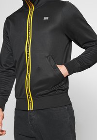 Sort zip-jakke med gul branding ned ad forsiden, med hætte og sidelommer. Materialet er glat med et tætsiddende design.