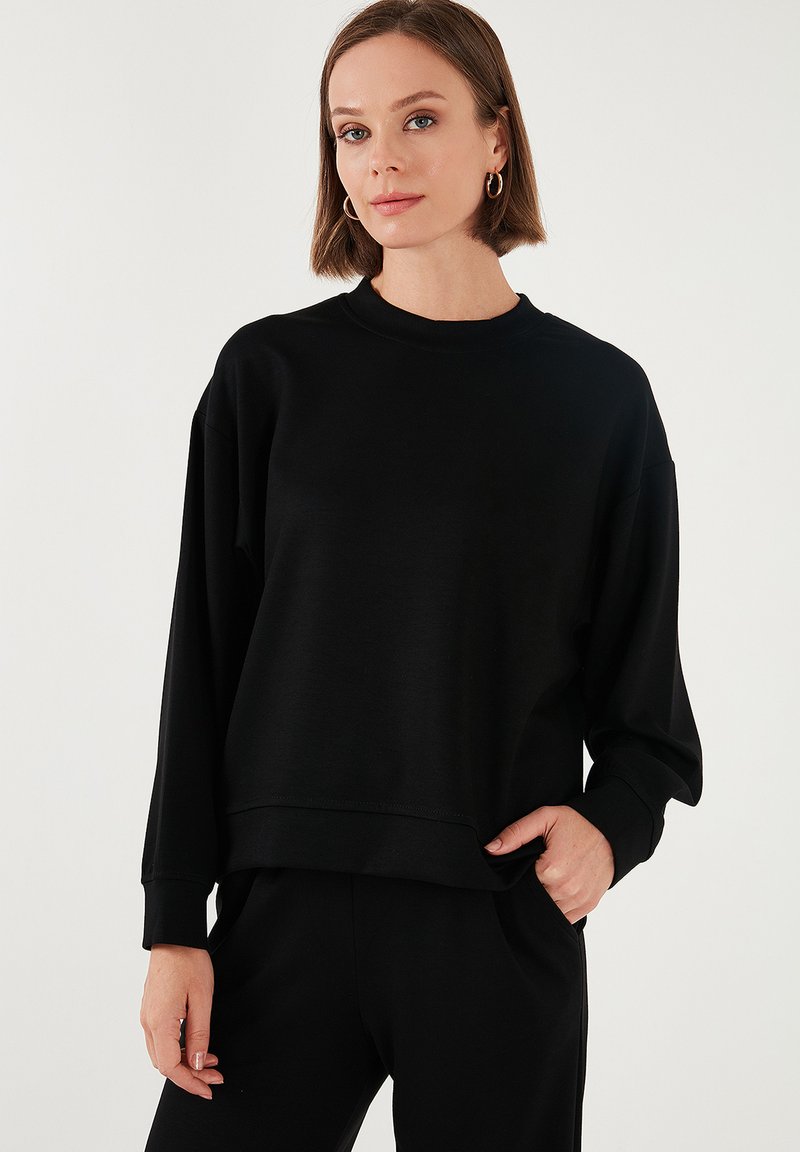 Felpa nera con maniche lunghe e larghe, colletto rotondo, texture morbida e polsini a costine, abbinata a pantaloni neri coordinati.