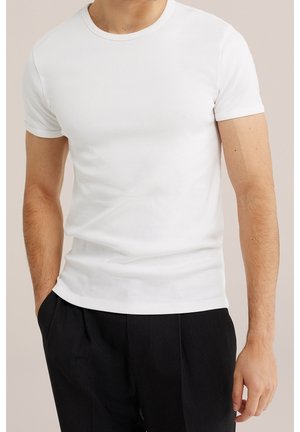 Mężczyzna noszący zwykły biały t-shirt z krótkim rękawem włożony do czarnych spodni, stoi z jedną ręką w kieszeni na neutralnym tle.