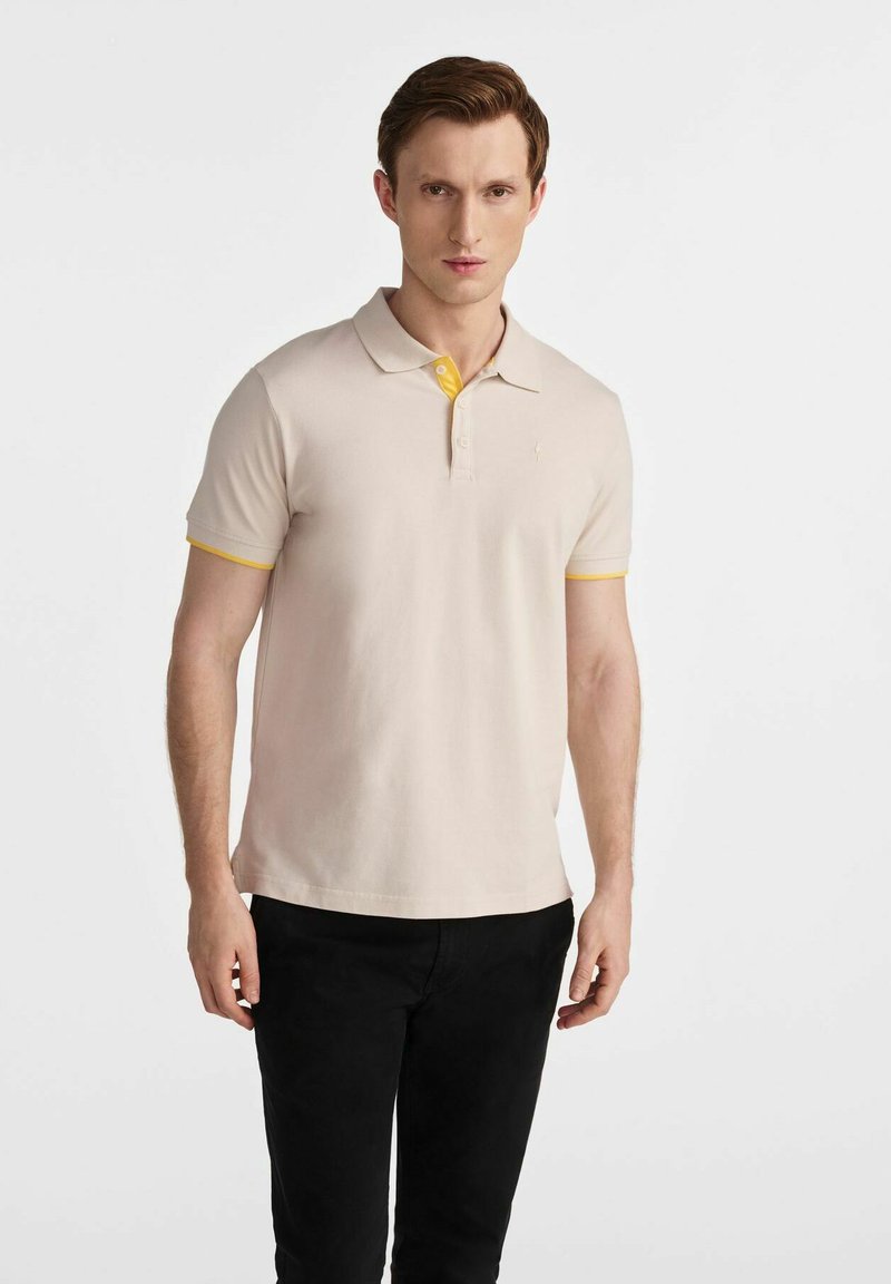 Beżowa koszulka polo z żółtym kołnierzykiem i akcentami na rękawach, z trzema guzikami i małym logo na piersi.