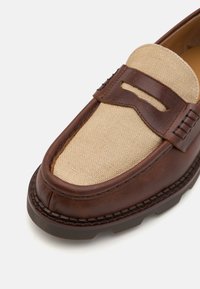 Mocassin en cuir marron avec une tige en toile beige. Les caractéristiques incluent un bout arrondi, des coutures décoratives et une semelle en caoutchouc pour une meilleure adhérence.