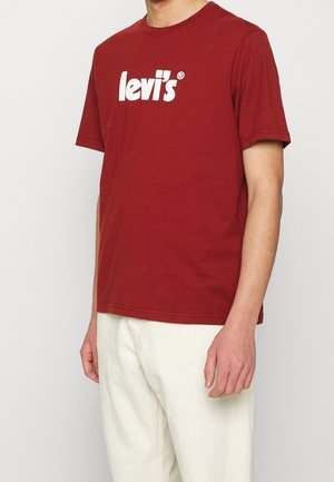 Man wearing a red Levi's t-shirt and off-white pants, standing against a plain light background.