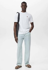 White short-sleeve t-shirt with a small logo, light blue jeans, dark shoulder bag, and brown sandals. Plain background.