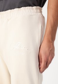 Beige Jogginghose mit elastischem Bund und Seitentaschen. Auf der Vorderseite befindet sich in erhabenem Druck der Text "Colucci la Famiglia".