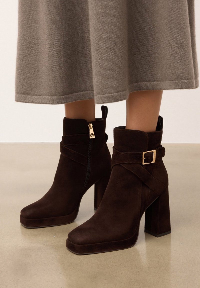 Bottines en daim marron avec talons carrés, fermeture éclair dorée et bride à boucle, portées avec un pantalon beige à jambes larges.