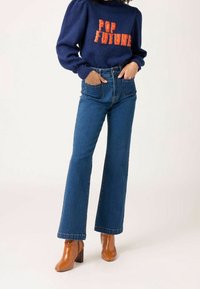 Pull bleu foncé avec le texte "POP FUTURE", porté avec un jean bleu taille haute et jambe droite. Des bottines marron à talons blocs complètent la tenue.