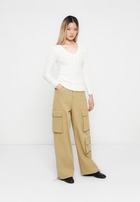 Sweater branco com nervuras em formato de V, combinado com calças cargo largas bejes que apresentam múltiplos bolsos. Modelo posando sobre um fundo simples.