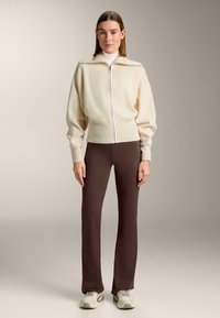 Roomkleurig fleecejack met ritssluiting en hoge kraag, gecombineerd met nauwsluitende bruine broek en lichtgekleurde sneakers. Zachte textuur, comfortabele pasvorm.