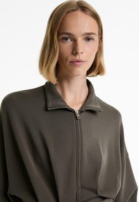 Olive green zip-up jacket with a high collar, ribbed texture, and a relaxed fit. Features a silver zipper hardware detail.