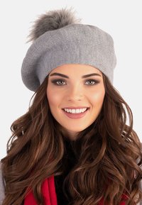 Chapeau béret en tricot gris avec un pompon en fourrure grise sur le dessus. Design simple avec une texture douce et un petit logo en accent.