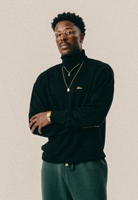 Άντρας με γυαλάκια, ντυμένος με μαύρο πουλόβερ τύπου τζέρσεϊ, χρυσές αλυσίδες, χρυσό ρολόι και γκρι παντελόνι, στέκεται με τα χέρια σταυρωμένα μπροστά σε απλό φόντο.