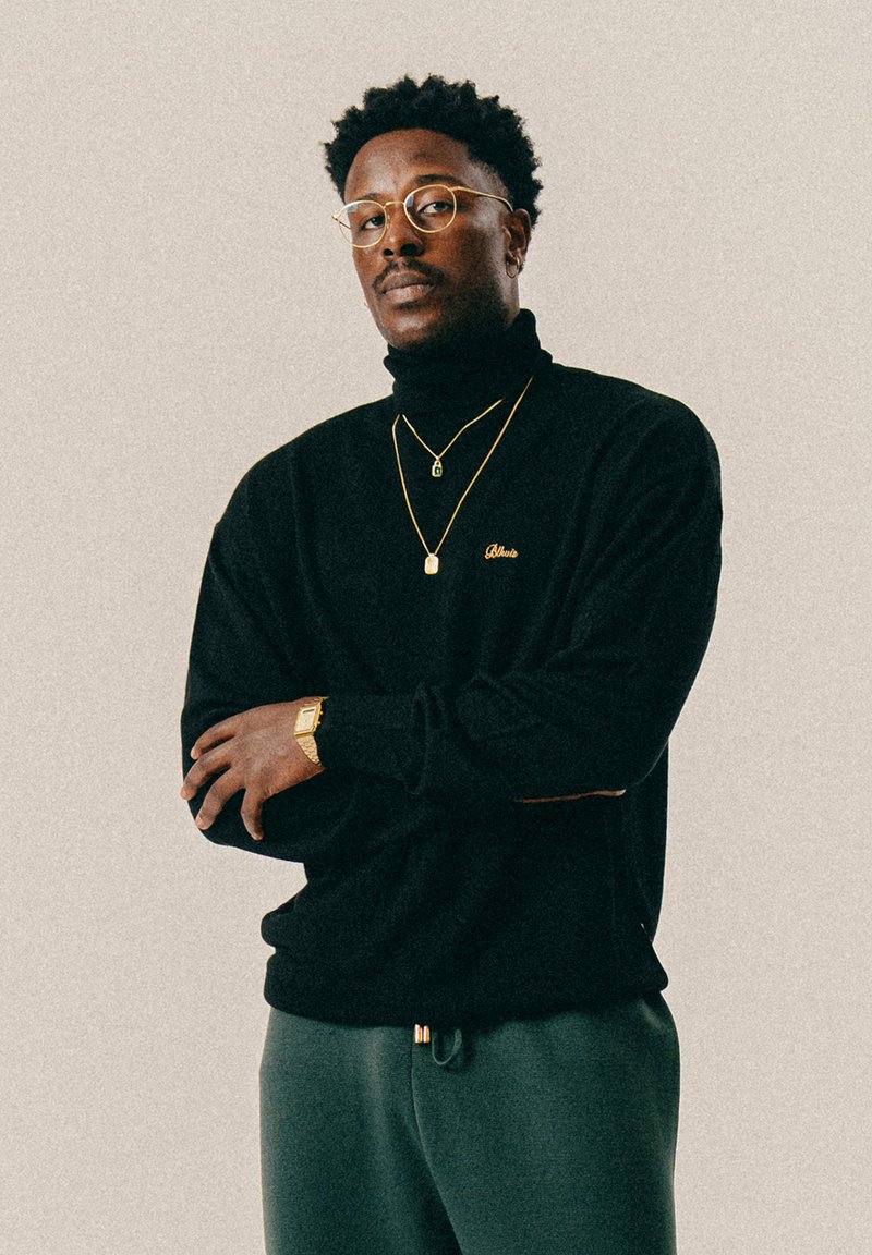 Άντρας με γυαλάκια, ντυμένος με μαύρο πουλόβερ τύπου τζέρσεϊ, χρυσές αλυσίδες, χρυσό ρολόι και γκρι παντελόνι, στέκεται με τα χέρια σταυρωμένα μπροστά σε απλό φόντο.