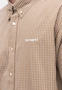 Καφέ και λευκό ριγέ πουκάμισο με κουμπιά και γιακά, με το λογότυπο της Carhartt κεντημένο σε λευκό στην αριστερή πλευρά του στήθους.