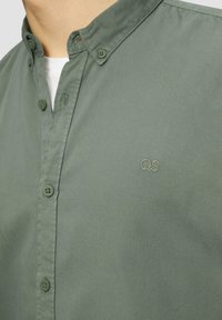 Groene overhemdbloes met een zachte textuur, puntige kraag en geborduurd "QS" logo op de borst. Regular fit met doorlopende knoopsluiting.