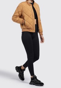 Femme portant une veste bomber matelassée beige, un crop top noir, des leggings noirs et des baskets noires épaisses, debout avec un pied en avant.