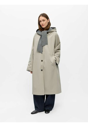 Femme portant un long manteau beige avec capuche et une écharpe à carreaux nouée autour du cou, pantalon large foncé et chaussures noires, debout la main dans la poche.