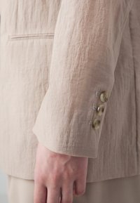 Manica di un blazer di lino beige con una texture sgualcita, caratterizzata da tre bottoni marrone chiaro e un sottile dettaglio di cuciture.