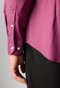 Gros plan sur le bras gauche d’une personne portant une chemise magenta à manches longues avec poignets boutonnés et un pantalon noir, devant un fond uni.