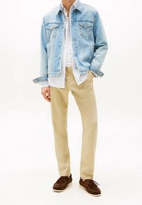 Hellblaues Jeansjackett mit zwei Brusttaschen, über einem weiß gestreiften Hemd getragen. Beigefarbene Hose und braune Wildlederboots vervollständigen das Outfit.