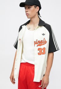 Kurzärmeliges Hemd mit Kragen in Weiß mit schwarzen Akzenten, versehen mit orangefarbenem Text und der Zahl 33. Kombiniert mit roten Shorts und einem weißen Tanktop.