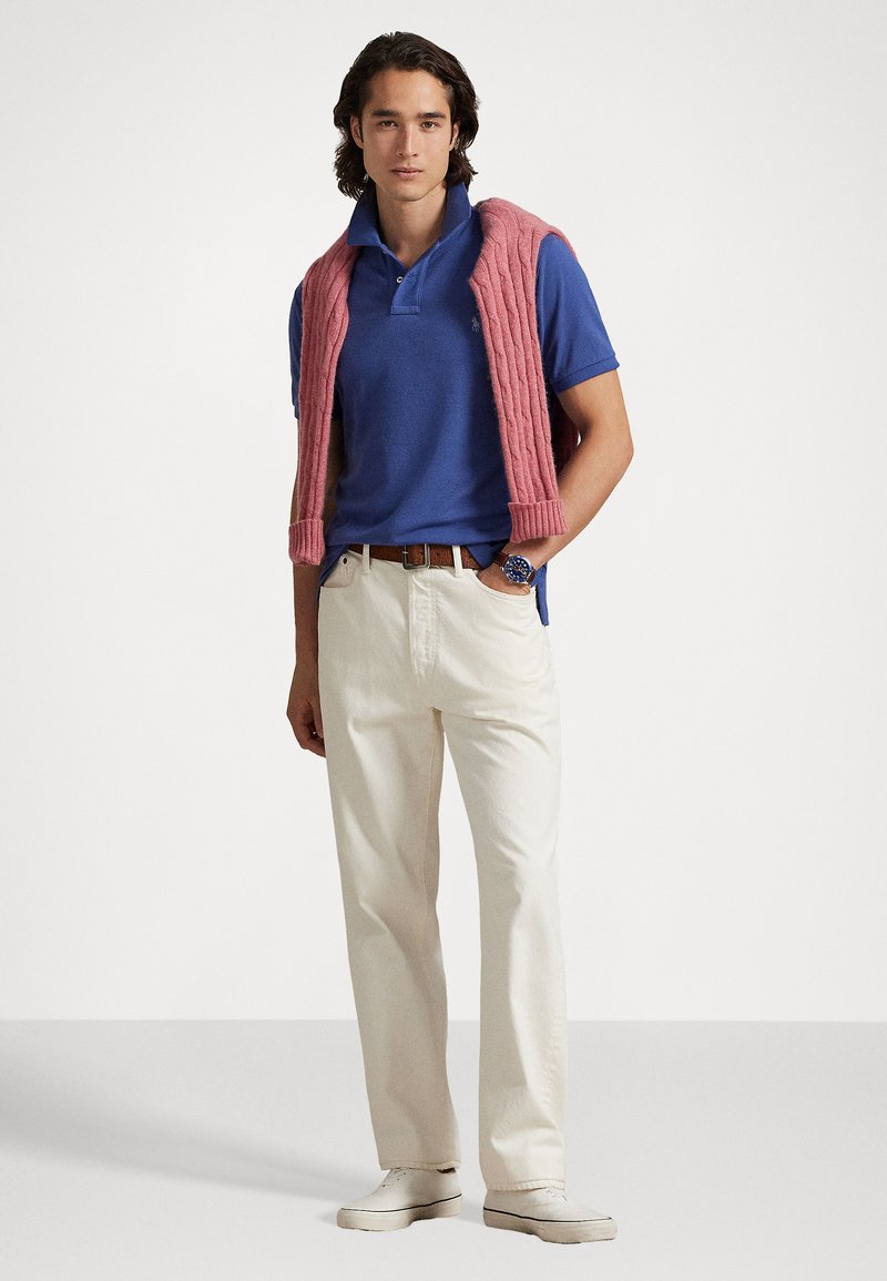 Blauwe polo, crèmekleurige broek, roze gebreide trui over de schouders, en witte schoenen. Opvallend horloge op de pols.