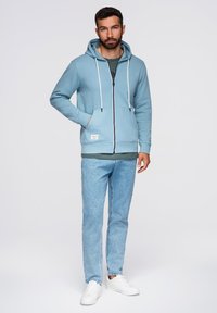 Jasnoniebieska bluza z zamkiem błyskawicznym i białymi sznurkami, zestawiona z zieloną koszulką i jasnoniebieskimi dżinsami, noszona z białymi trampkami.