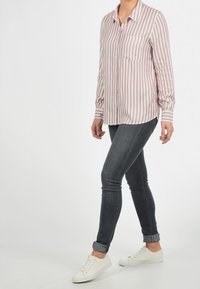 Donna che indossa una camicia a righe verticali bianche e rosa, jeans skinny grigi scuri arrotolati alla caviglia e sneakers bianche, in piedi su uno sfondo bianco.