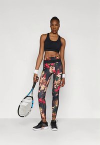 Žena u crnom sportskom grudnjaku, tajicama s cvjetnim uzorkom i tenisicama, drži tenisku reketa i stoji uz jednoličnu pozadinu.