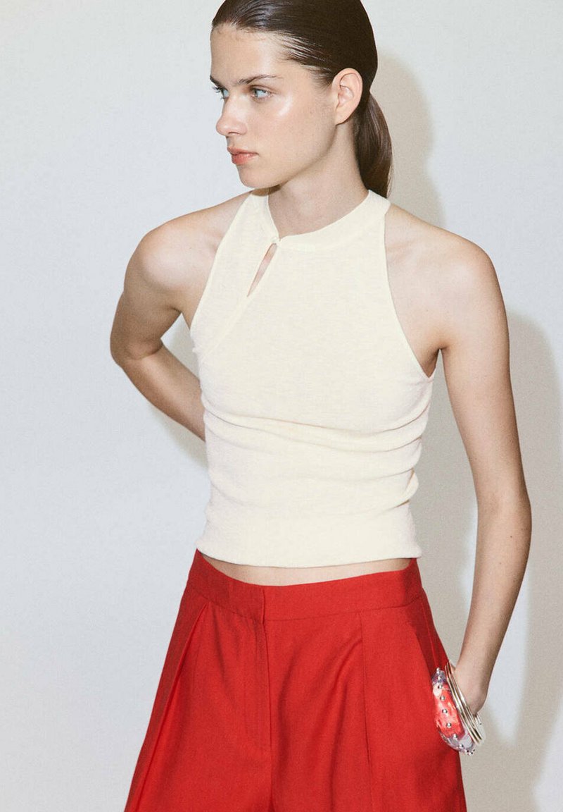 Jeune femme aux cheveux foncés portant un haut blanc sans manches et un pantalon rouge taille haute, regardant sur le côté avec la main dans la poche.